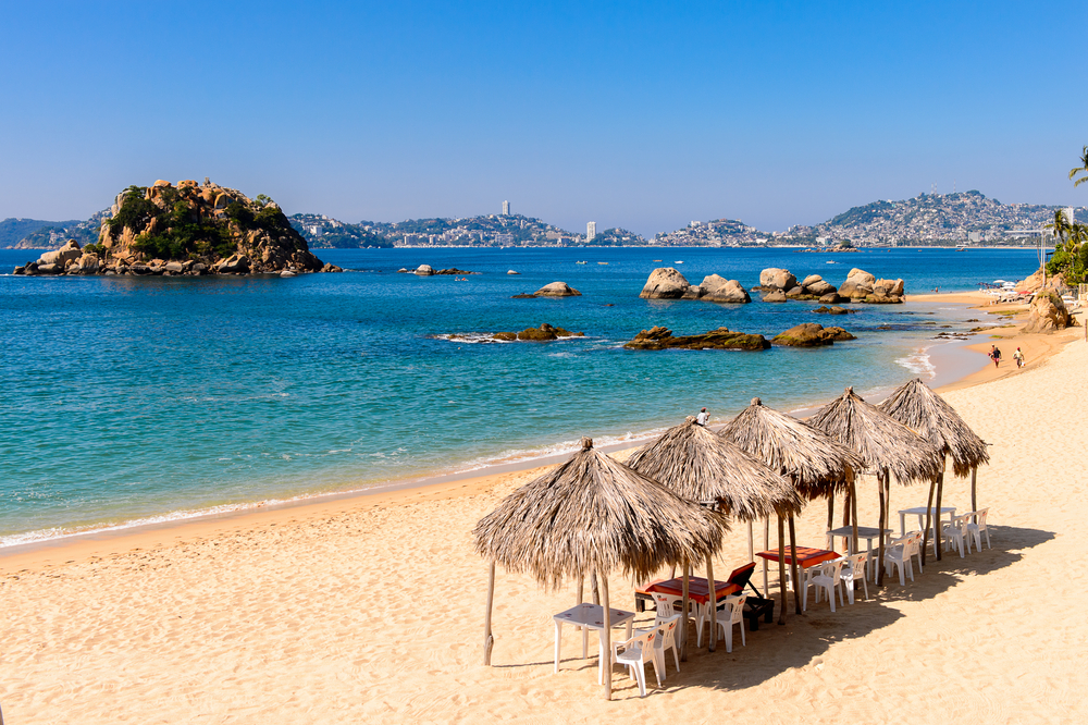 A beautiful beach with native umbrellas where a few people are seen walking on the shore.