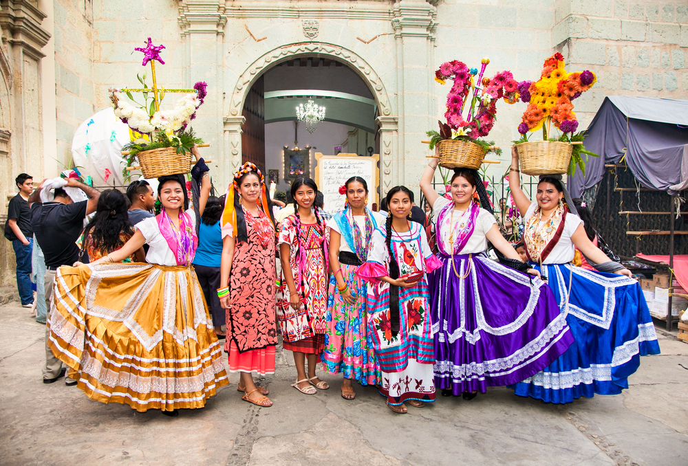 Latina women wearing vibrant festival clothes and others are carrying basket filled with flowers during a festival. 