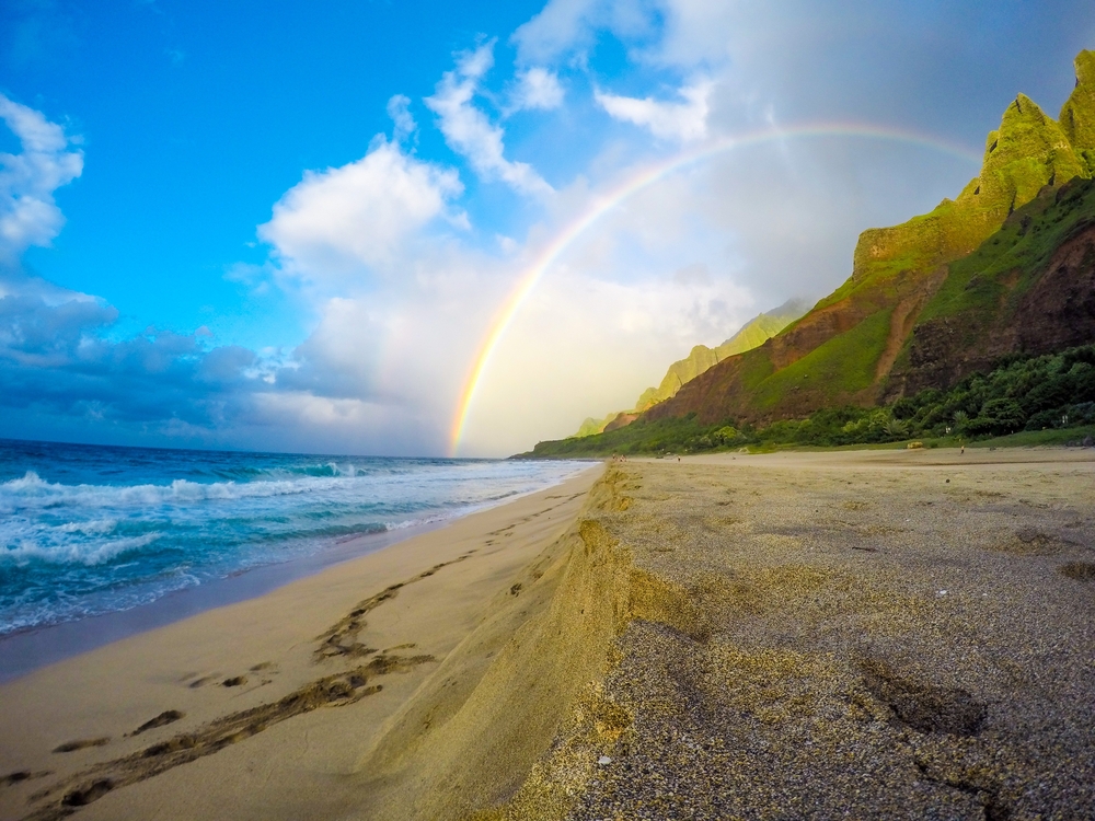 Beach view from the Napali Coast in Kauai, Hawaii with a rainbow arching over the sand and volcanic mountains for a piece answering what language is spoken in Hawaii
