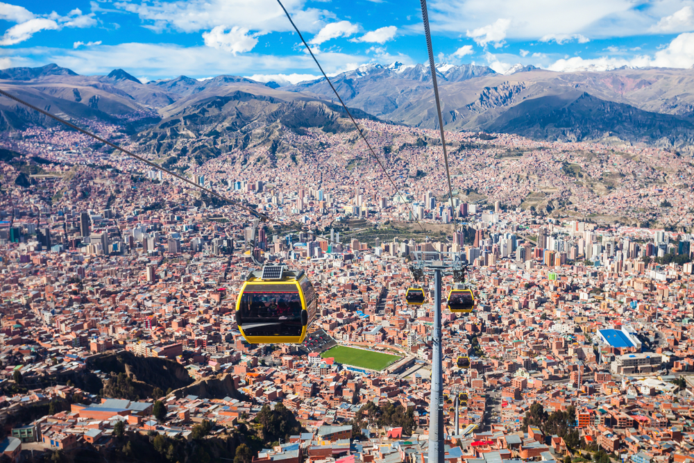 Cable cars traversing above a very populated city surrounded by tall mountains. 