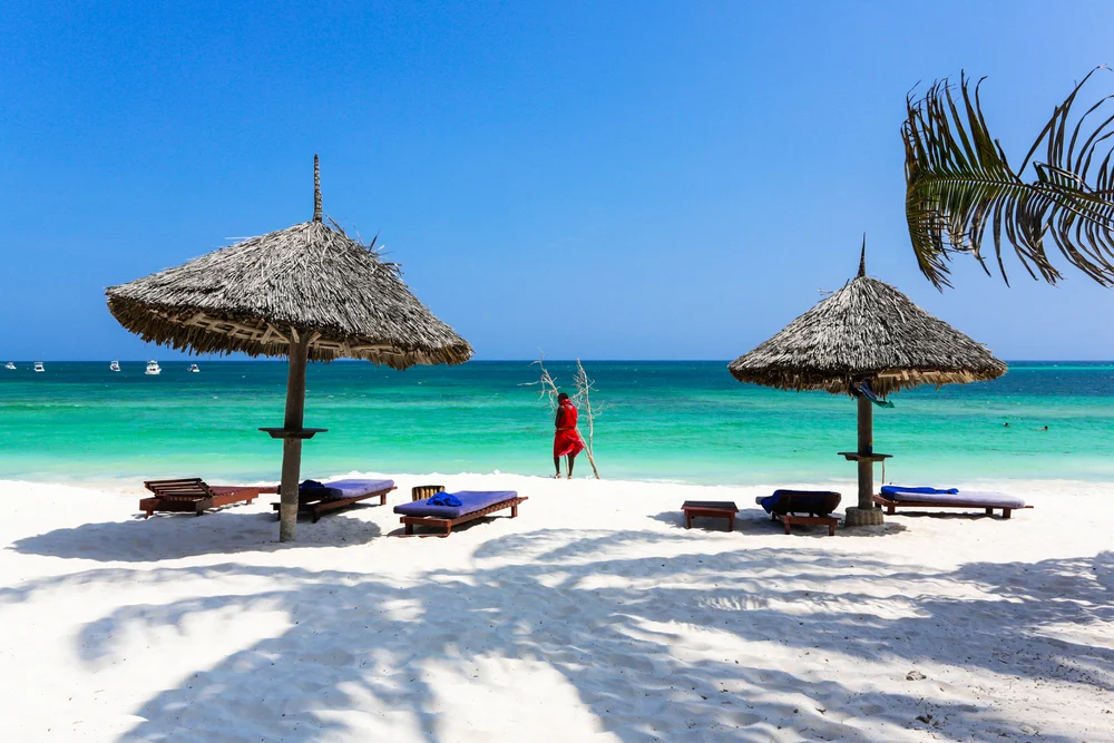 A man wearing red standing on the shore while facing the beach at Watamu, one of our picks on the best areas to stay in Kenya, six sun beds lie under two native umbrellas. 