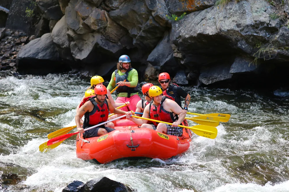 A group of friends white water rafting using a red raft while each of them are holding oars around a rocky river.