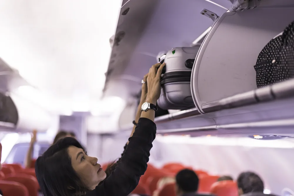 Female traveler loads a bag into the overhead bin in a plane cabin before flying for a guide showing how much Frontier baggage fees are 