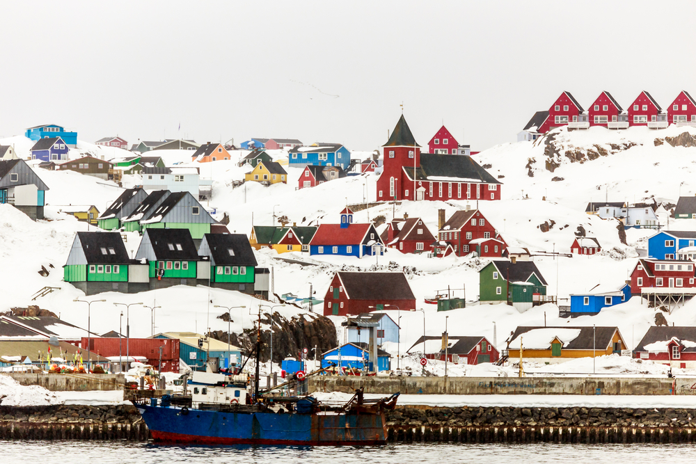 A coastal town on a hill covered with snow in Sisimiut, one of our picks on the best areas to stay in Greenland, and a boat is docked on the pier.