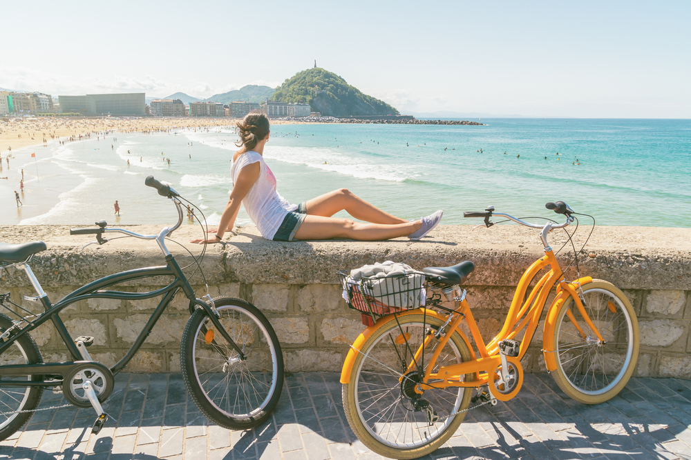 A woman enjoying the view of a beach while seating on a concrete near two bicycles, looking at the people enjoying the shore.
