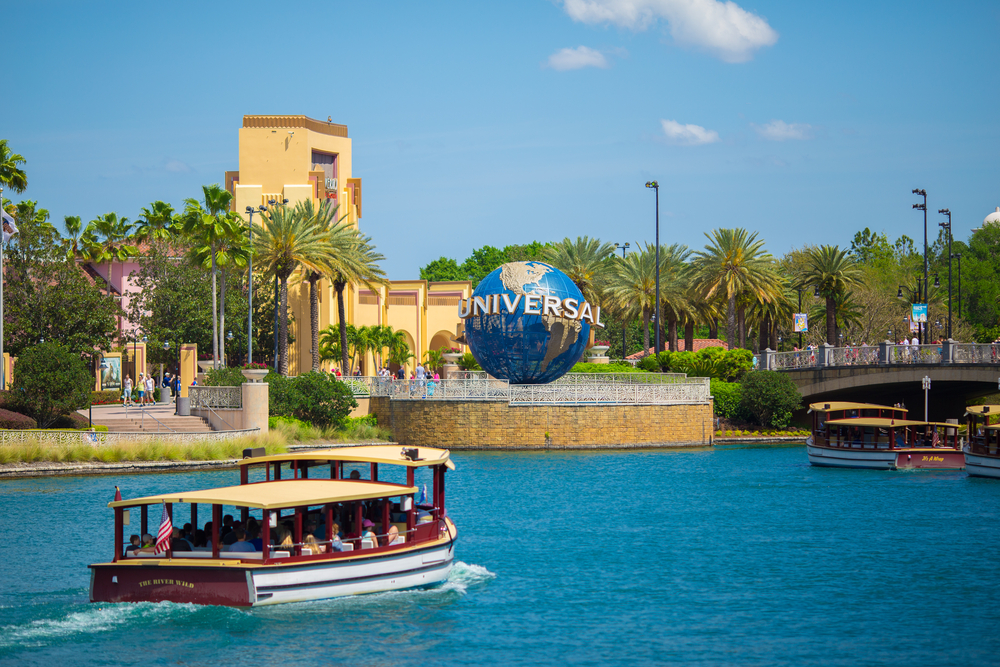 A small boat touring on the river at the Universal Studios Florida, an image for an article about trip cost to Orlando, the large Universal Studio globe figure is beside the river.