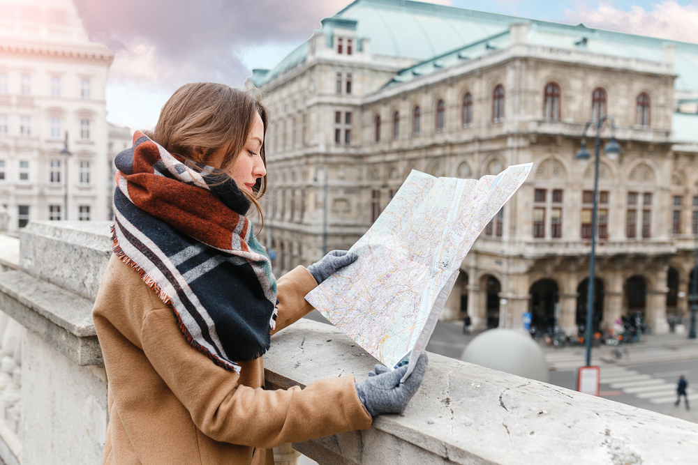 A woman wearing scarf, coat, and gloves while holding a map of a city, a concept image for a travel article about safety in visiting Vienna.