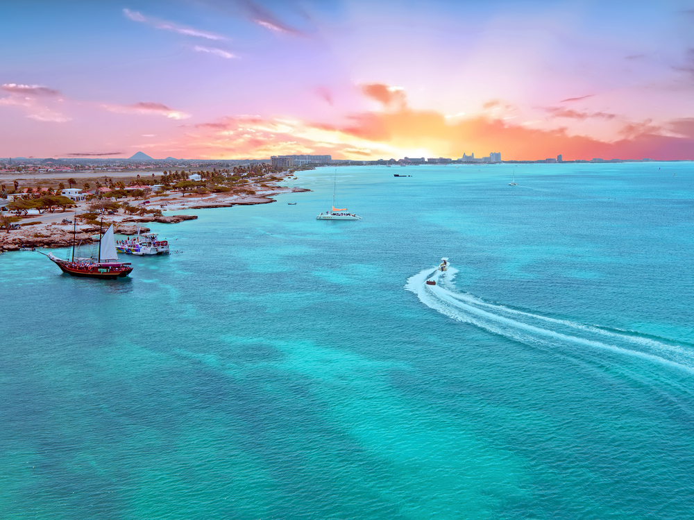 Aerial view of the island of Aruba at sunset with brilliant colors in the sky as a boat moves through the water, showing the concept of how long is a flight to Aruba for an article answering the question