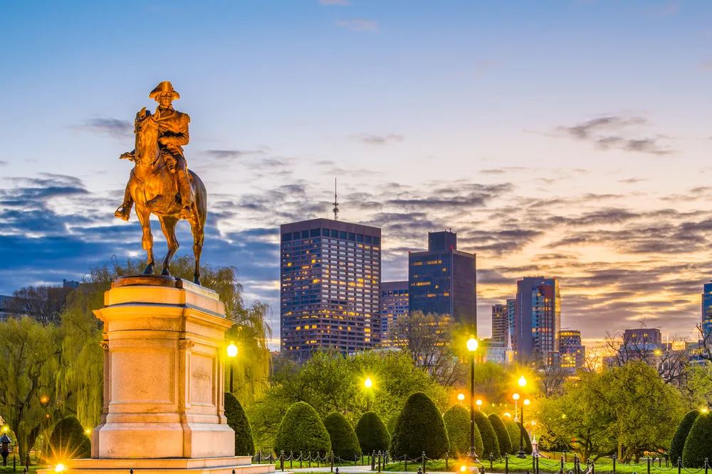 A statue of a man riding a horse at the center of a park with well-trimmed trees, photographed during a sunset for a piece on a travel guide about trip cost to Boston.