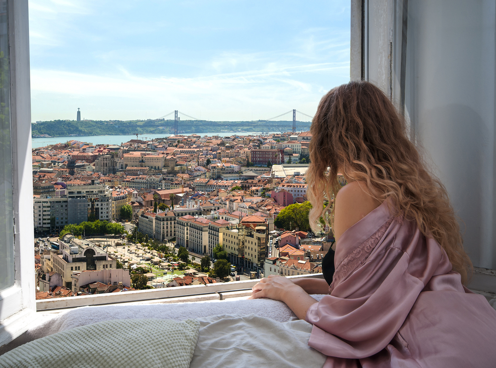 A woman in a hotel room looking at the window with the view of a city and a long bridge.