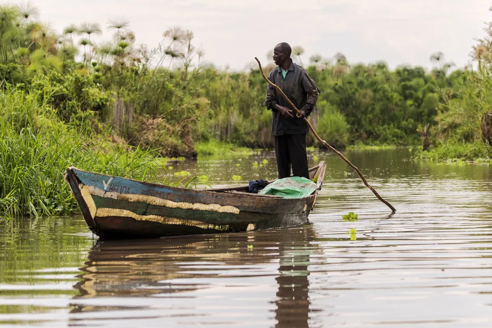 A man standing on his small fishing boat while pushing his boat using a long piece of wood. 