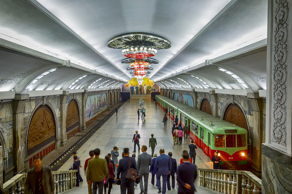 Several people walking down a subway station with a chandelier and on a stand by green-red train. 