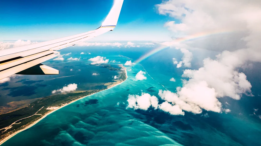 Close-up view of an airplane wing and a rainbow as it flies over a tropical island to show the concept of how long is a flight to Hawaii