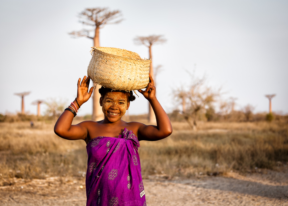 A local wearing pink while carrying a straw basket over her head, smiling for the camera, and the Baobab trees in background. 