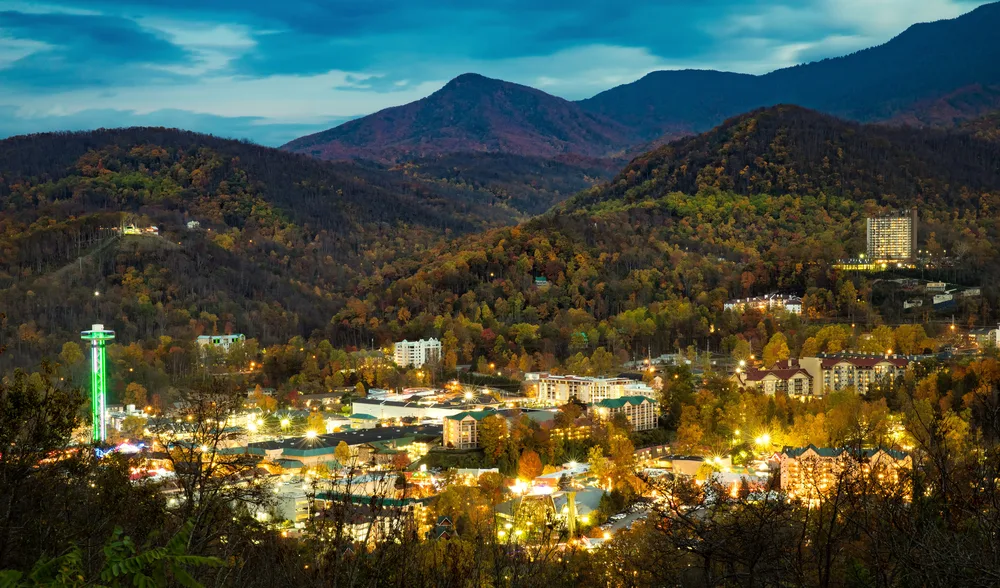 A small town lit during dusk, surrounded by lush trees and towering mountains, an image for an article about trip cost to Gatlinburg.