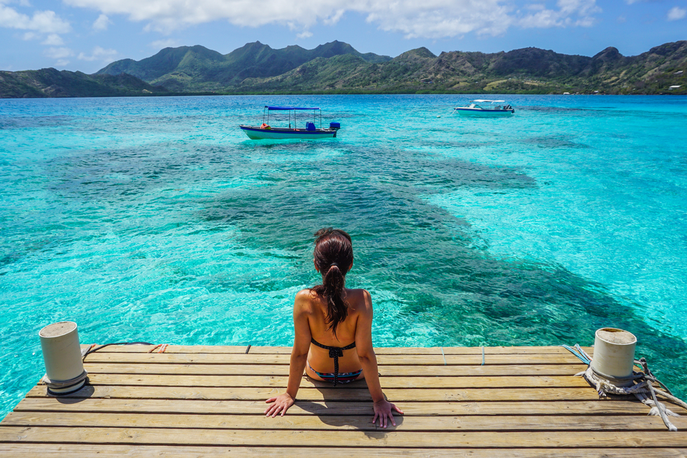 A woman seating on a dock at the beach while looking at the boat ahead in a crystal clear water. 