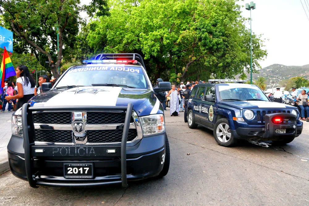 Police cars parked on the side of the street while a large crowd is at the back during a local celebration. 