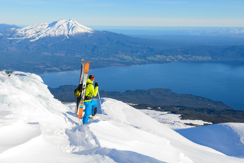 A man wearing yellow and blue preparing to ski down from the peak of a snow-covered mountain , and another tall mountain can be seen from a distance with snow cap, an image for an article about trip cost to Chile.
