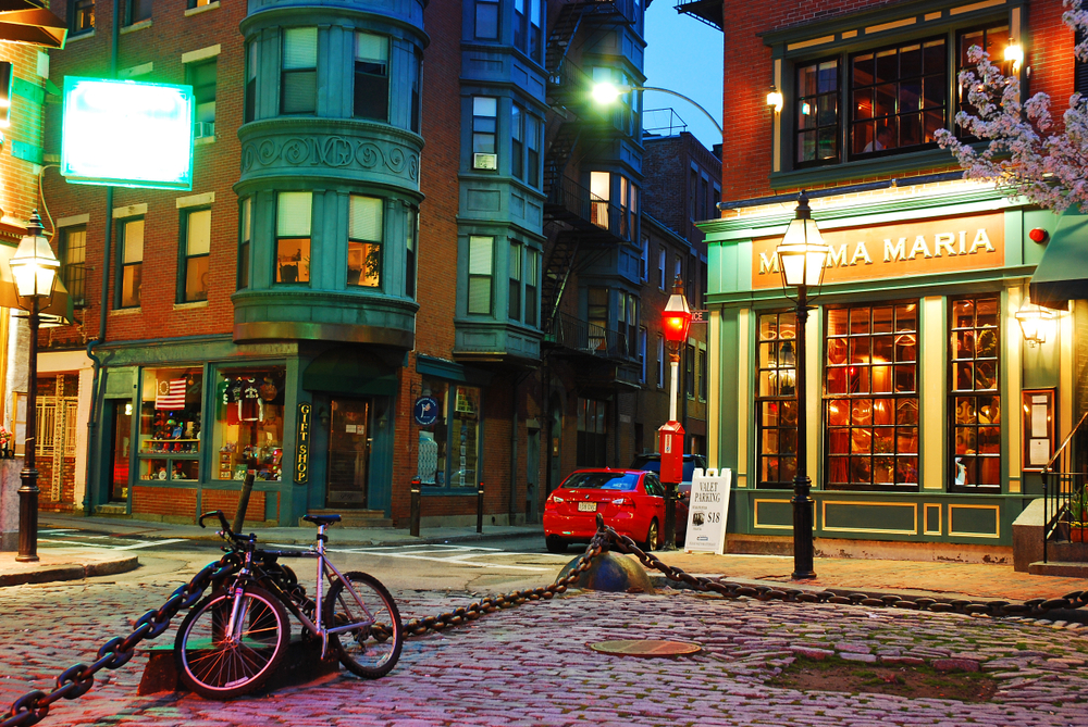 Bike chained to a fence at dusk in the Boston North End Italian neighborhood with colorful, lit shops around the cobblestone streets for a guide to spending a weekend in Boston
