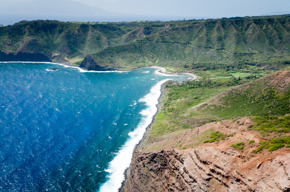 Aerial view of Molokai's coastline with rugged mountains and sand next to terraced, weathered ridges by the ocean for a guide showing how many people live in Hawaii's Molokai and other islands