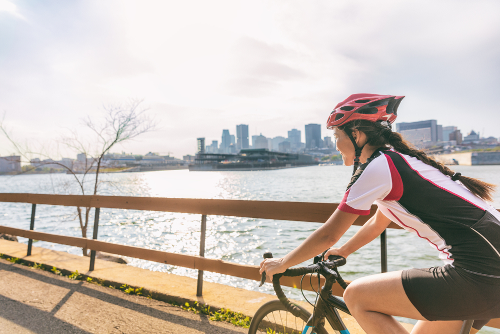 A woman cycling by the riverbank while wearing a cycling helmet and buildings can be seen on the other side of the river, captured during the best time to visit Montreal.
