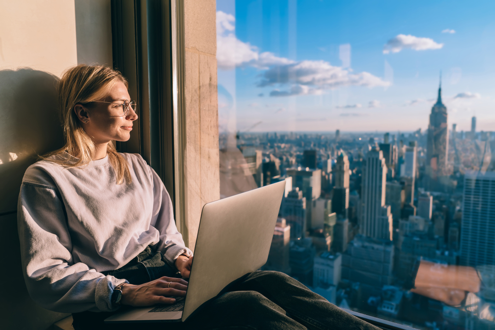 A woman sits by her hotel window looking out at the New York City skyline as she uses her laptop for a work trip to indicate the concept of bleisure, or traveling for both business and leisure