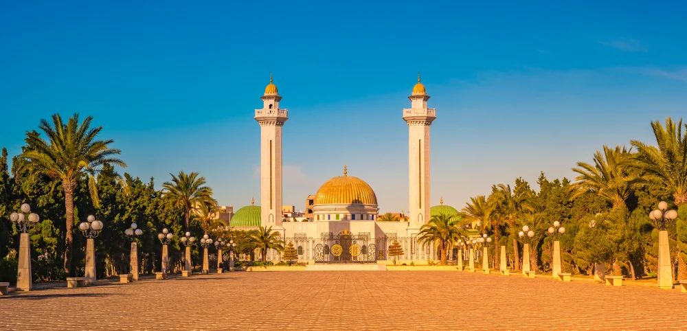 A rich mosque with a vast open grounds paved with bricks and some areas are planted with palm trees. 