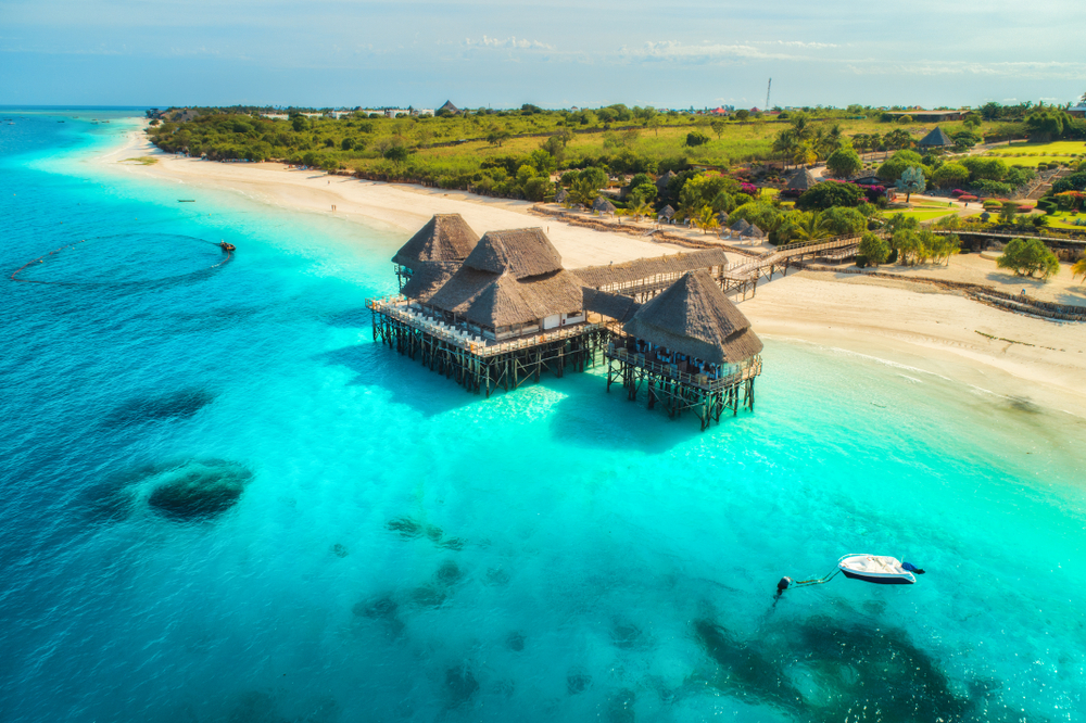 Aerial view on a majestic beach with crystal clear water and a native hut built on the beach that has a footbridge that leads to the land. 