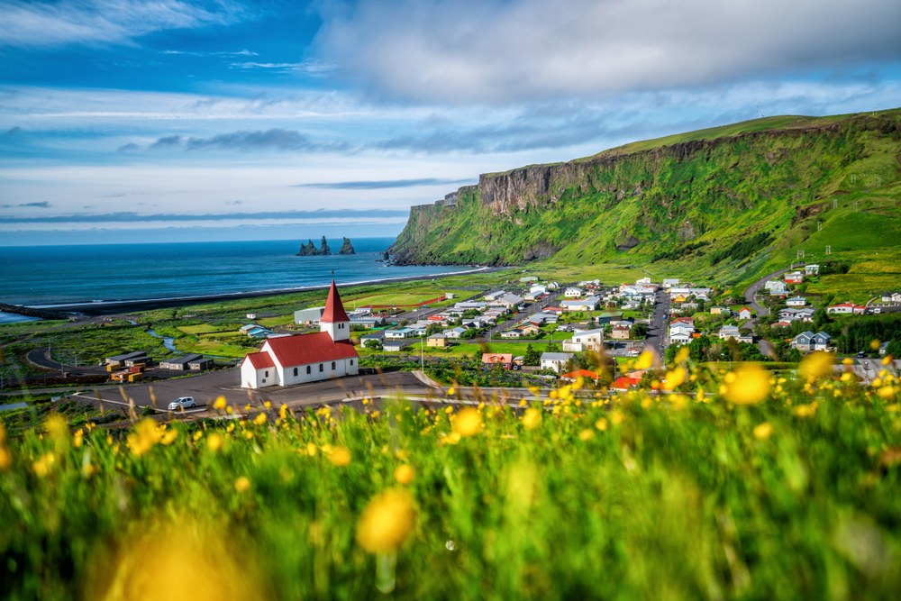 Iceland's southernmost village, Vik I Myrdal, in summertime with yellow wildflowers and red-roofed church surrounded by homes and buildings by the sea for a guide explaining how long flights to Iceland take