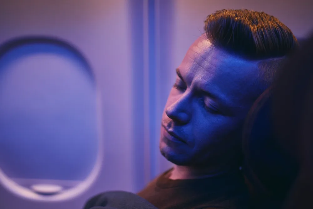 Man sleeping on an airplane during a red eye flight with soft, purple-colored light around him 