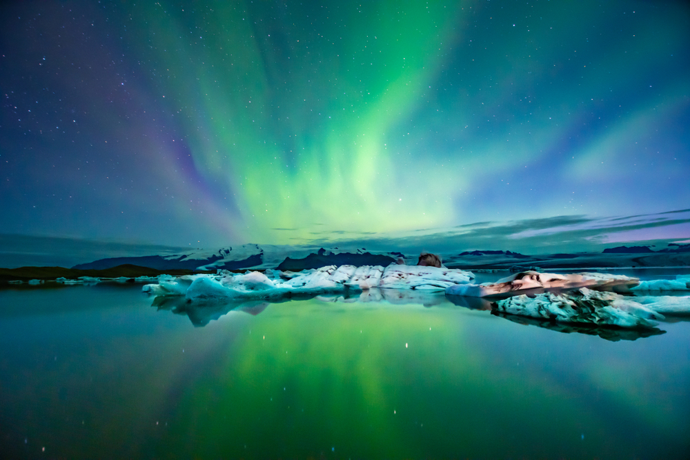 The Glacier Lagoon in Iceland seen with green and purple Northern Lights in the skies above at nighttime for a guide exploring how long is a flight to Iceland from the US