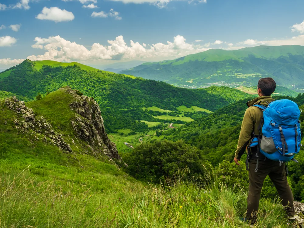 A hiker carrying a loaded blue backpack standing on the side of a mountain while looking ahead the green mountains during the overall best time to visit Armenia.