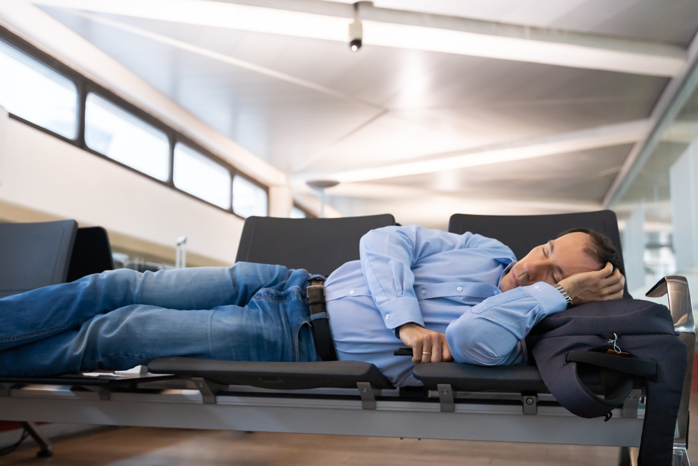 Man lays across several seats at the airport to show the difference between a layover vs stopover while he waits for a connecting flight