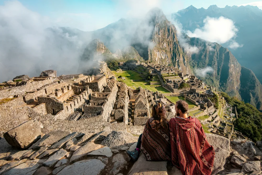 A couple wearing native cloaks while seating on a rock overlooking a ruins of an ancient structures at the peak of a mountain, a piece on the guide on facts about South America.