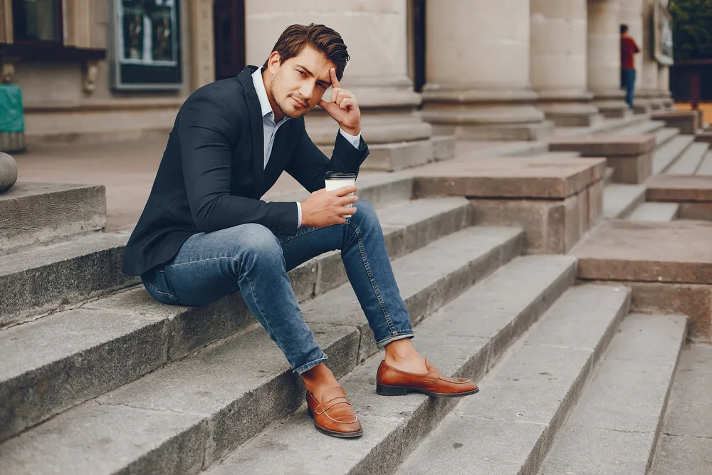 A man wearing a suit paired with jeans, as he seats on the steps. 