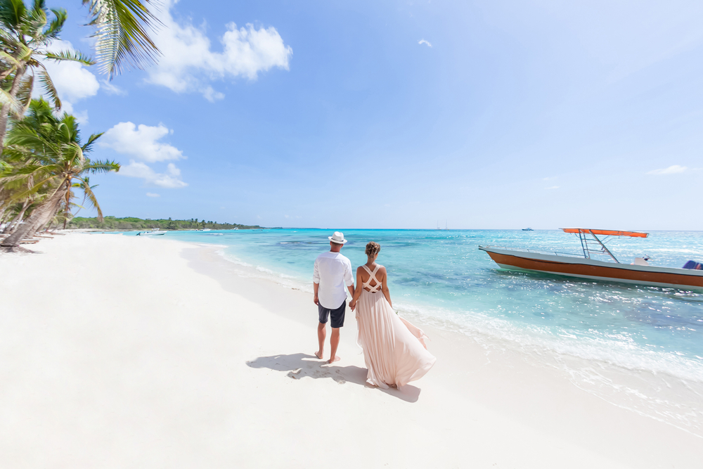 A couple walking on a white sand shore where a small boat can be seen in the waters.
