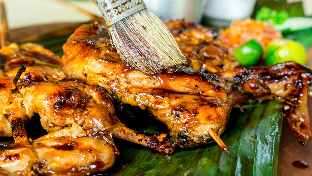 Applying sauce using a brush to a grilled chicken prepared on a banana leaf, for an image to the food, drinks, and activity cost on an article about trip cost to the Philippines.