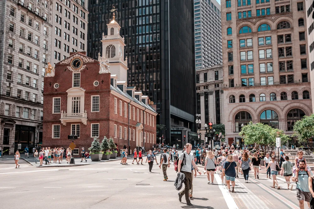People walking on a square where an old buildings can be seen surrounded by modern buildings, an image for a guide on the best time to visit Boston.
