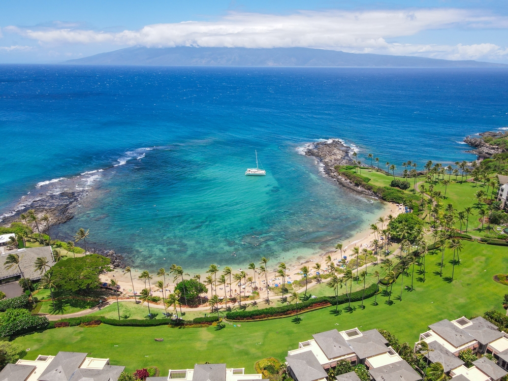 Aerial view of the Kapalua Coast in Maui with a boat in the water and crescent-shaped beach curving around on a clear day for a piece explaining the airlines that fly to Hawaii