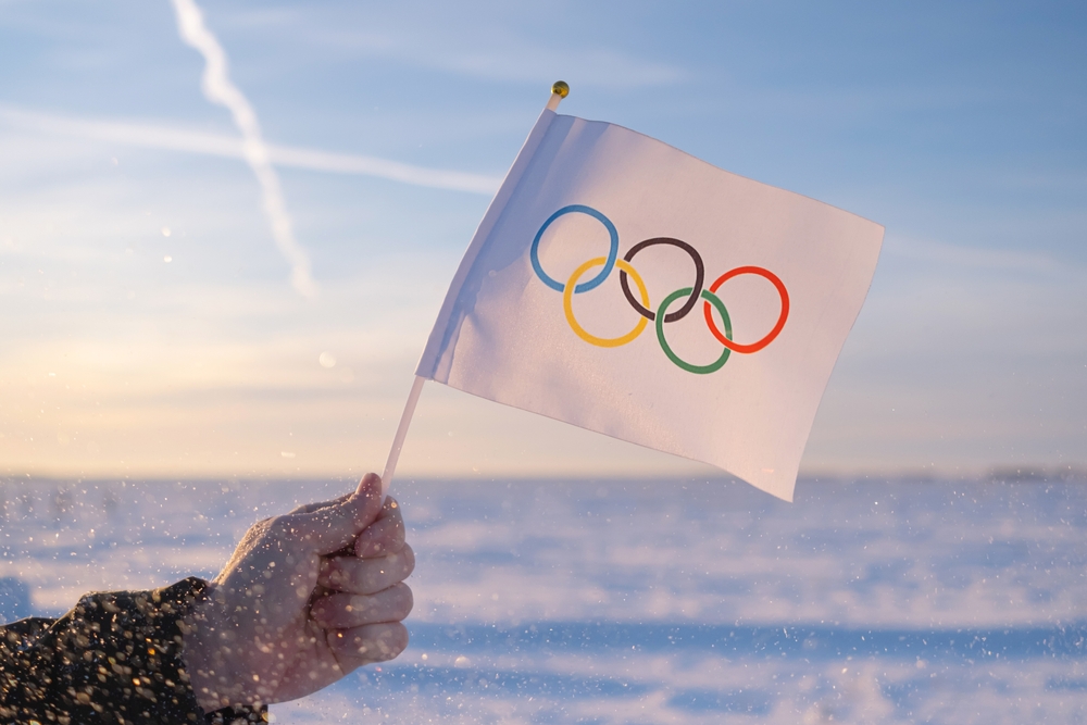 A small olympic flag waved by a person with a background of the sea. 
