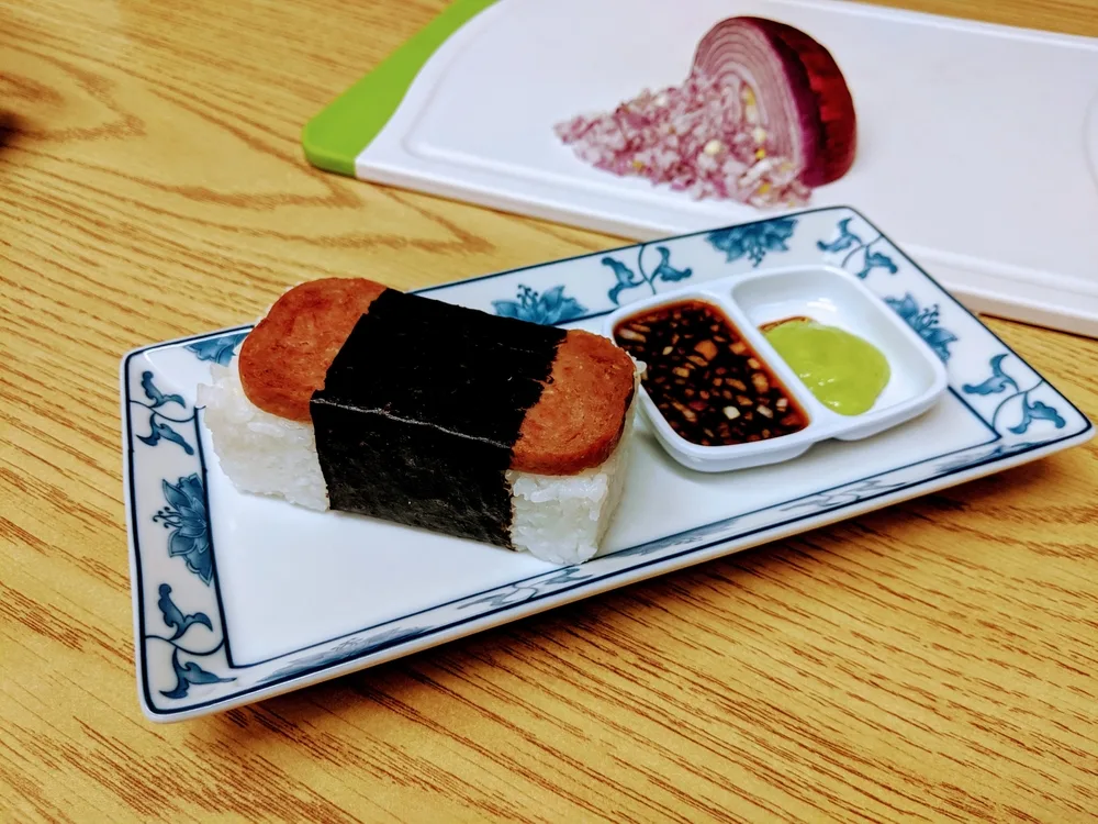 SPAM musubi on a rectangular plate with dipping sauces and red onion on a cutting board for an article answering why is Spam so popular in Hawaii?