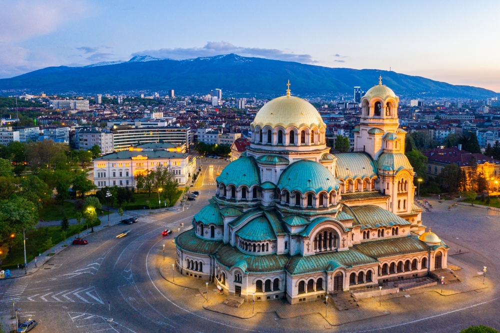Aerial view of an old church at the center of a roundabout, and a city and a tall mountain is visible in background, for a piece on what is in Eastern Europe.