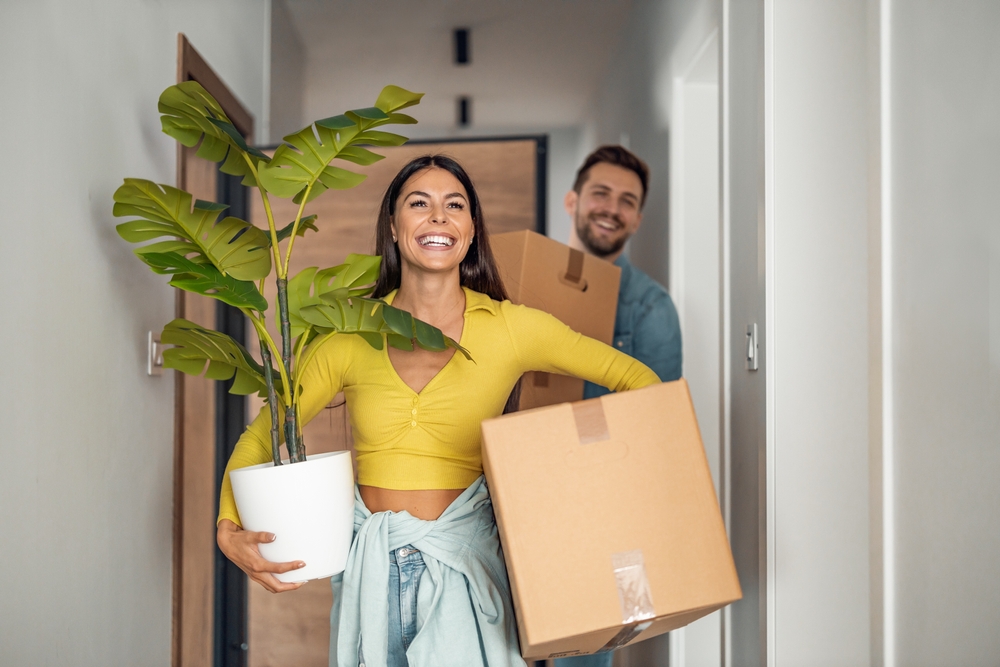 Young couple smiles as they carry boxes and houseplant into their new home to show the concept of how to move to Hawaii