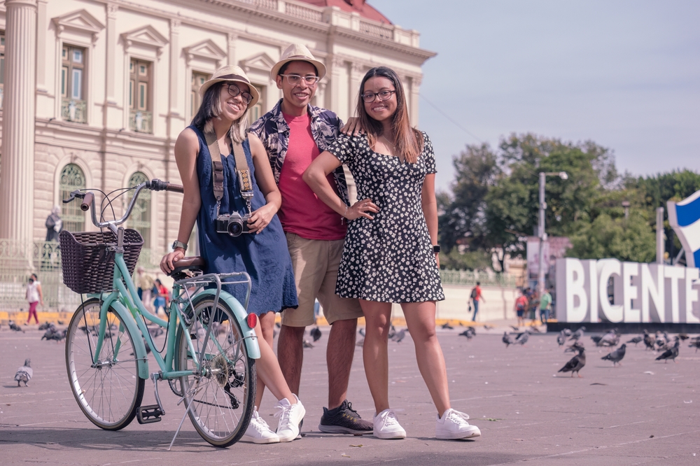 Three friends posing for a photo, where one person can be seen leaning on a bike, and pigeons can be seen in background, an image for the fact about Salvadorians living in cities.