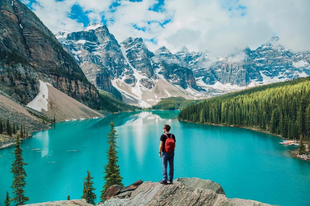 A man standing on a rock facing a calm lake surrounded by icy mountains and forest at the Banff National Park, an image on the guide about the best time to visit the area.