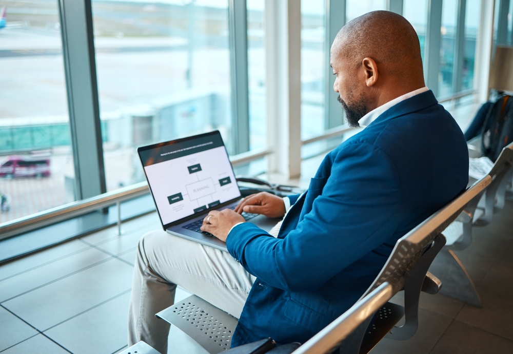 African American man sitting in an airport to show the concept of what is a layover as he works on his laptop wearing a business suit