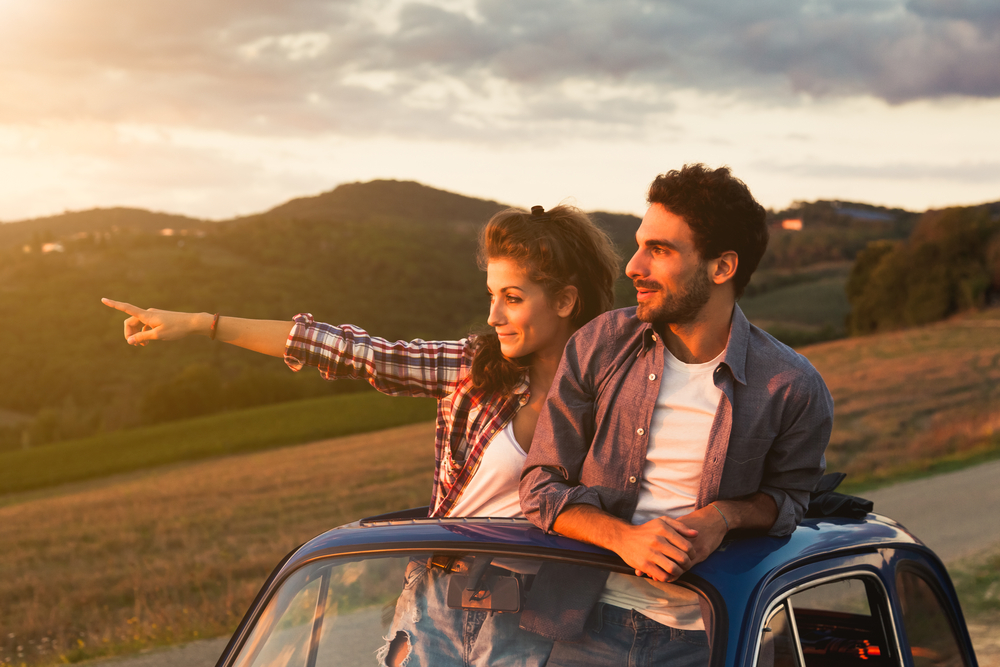 A couple watching the sunset where the woman is pointing towards a certain direction, while the top half of their body is out is the car's sunroof. 