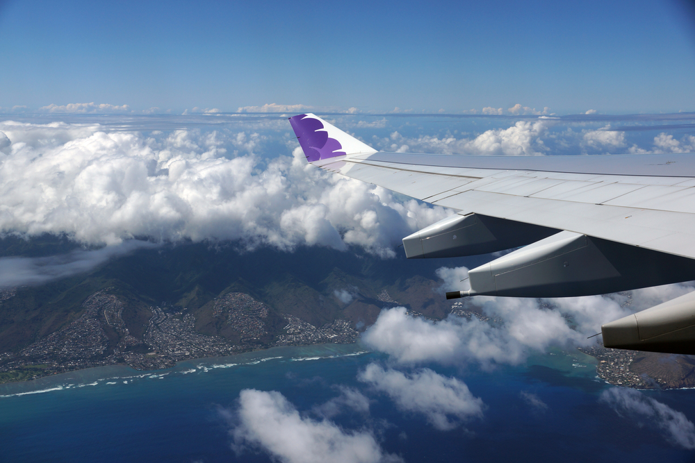 Hawaiian Airlines plane wing tipped in purple with clouds underneath as it passes over Hawaii for a guide showing which airlines fly into Hawaii