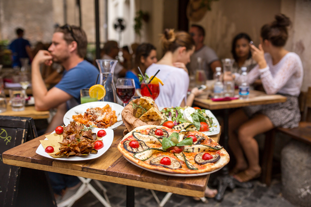 Pizza pies and other food prepare on a small wooden table, where people are blurred in background. 