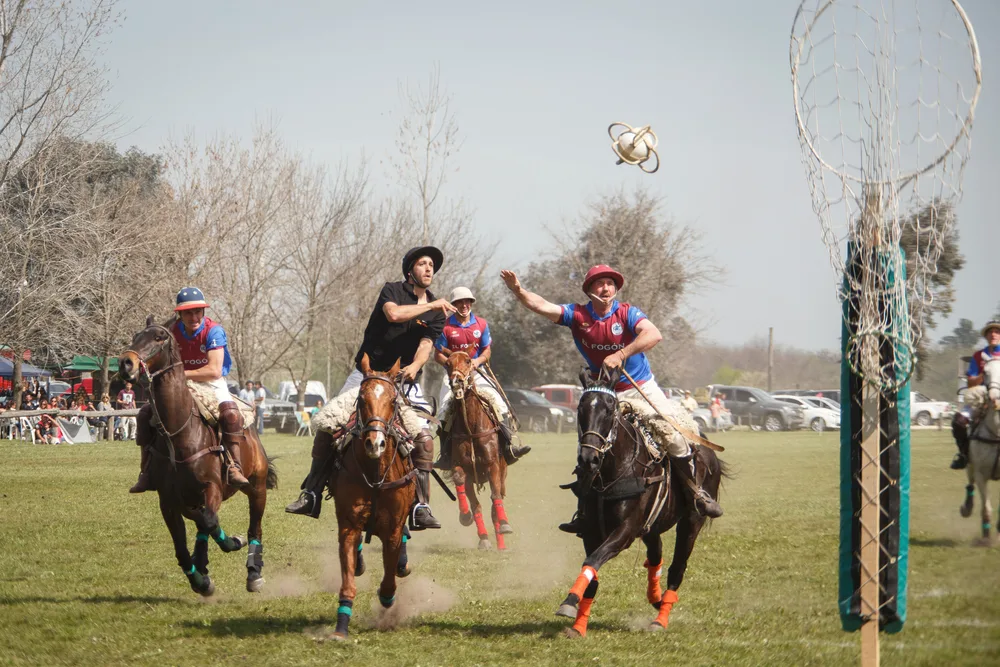Men playing a sport where they are mounted on horses and they are trying to shoot a ball in a vertical hoop. 